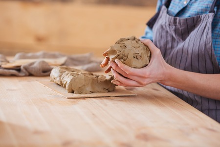 Closeup of piece of clay holded by hands of woman potter in apron sitting at the wooden tableの写真素材