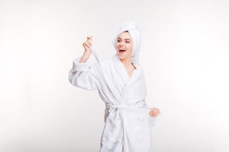 Cheerful joyful young woman in bathrobe with towel on her head standing over white backgroundの写真素材