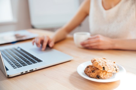 Closeup of young businesswoman using laptop and drinking coffee with cookies on workplaceの写真素材