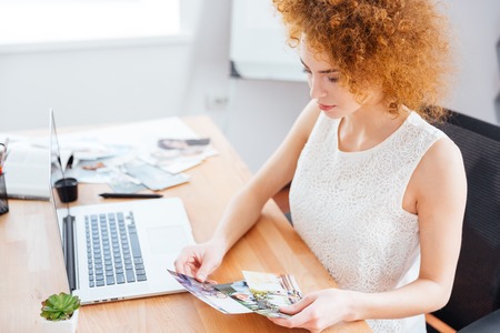 Cute lovely redhead young woman photographer sitting on workplace and choosing photosの写真素材
