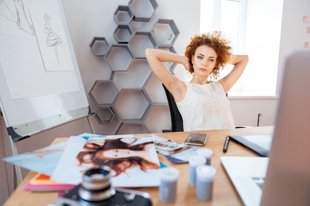 Relaxed pensive curly young woman photographer sitting on workplace with hands behind head