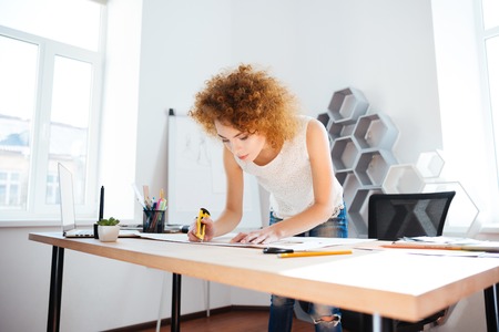Serious attractive young woman photographer with curly red hair working in office using stationery...