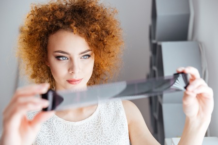 Closeup of attractive curly young female photographer holding and looking at photographic film