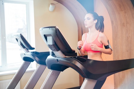 Pretty focused young woman athlete in earphones running on treadmill in gym