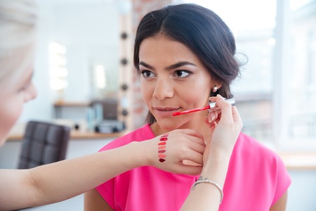 Female makeup artist testing lip gloss on hand and choosing perfect color for beautiful young womanの写真素材