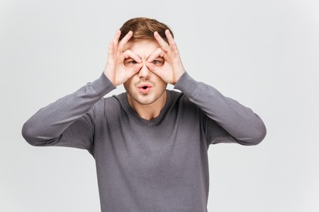 Funny young man in grey pullover making glasses with hands over white backgroundの写真素材