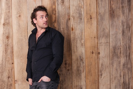 Attractive curly young man in black shirt standing with hands in pockets and looking up over wooden backgroundの写真素材