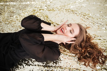 Happy excited young woman with long curly hair lying with eyes closed over sparkling backgroundの写真素材