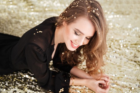 Cheerful beautiful young woman in black dress with curly long hair lying over shining backgroundの写真素材