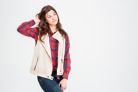 Pensive pretty young woman in plaid shirt and waistcoat standing and touching her hair over white...