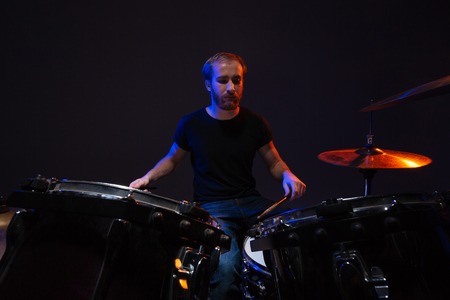 Attractive concentrated bearded man drummer playing his kit over dark background