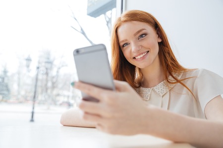 Cheerful charming young woman with long red hair using smartphone in cafeの写真素材
