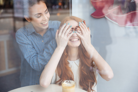 Smiling attractive young woman covered eyes by hands to cheerful redhead girl sitting in cafeの写真素材