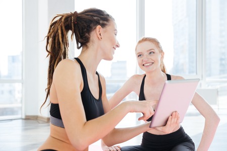 Two cheerful young sportswomen smiling and using tablet togetherの写真素材