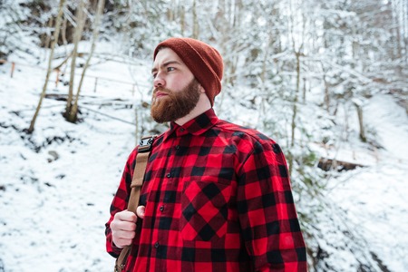 Portrait of serious bearded young man in chekecred shirt and hat with backpack in winter mountain forestの写真素材