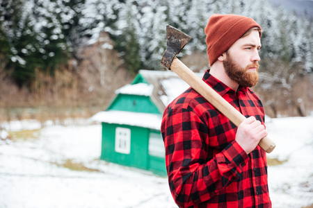 Handsome young man in checkered shirt and hat with axe walking in villageの写真素材