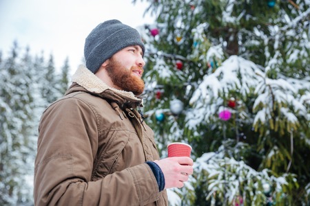 Thoughtful smiling bearded young man standing near decorated christmas tree and drinking coffee outdoorsの写真素材