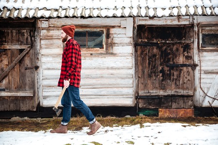 Handsome bearded man in checkered shire and jeans holding axe and walking in villageの写真素材