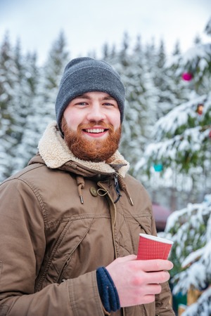 Happy young bearded man drinking hot coffee near decorated christmas tree in winter forestの写真素材