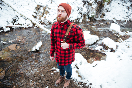 Top view of pensive bearded young man in checkered shirt and hat standing near mountain river in winterの写真素材