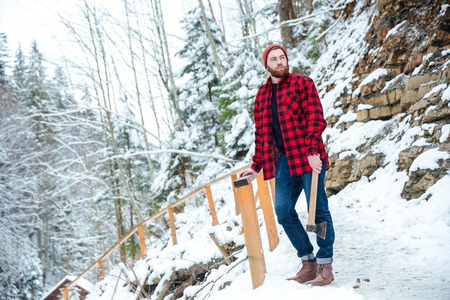 Thoughtful attractive young man with beard standing and holding axe in mountains in winterの写真素材