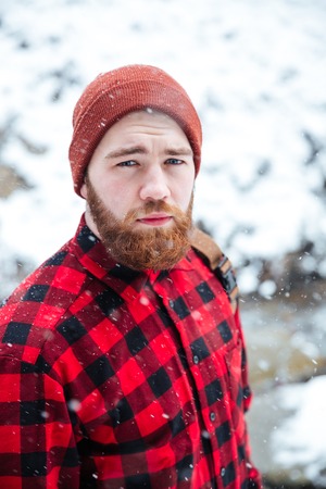 Portrait of serious handsome bearded man in checkeres shirt and hat outdoors in snowy weatherの写真素材