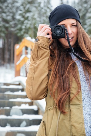 Cheerful pretty young woman with long hair taking photos of you on stairs in winter forest