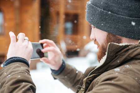 Concentrated bearded man taking pictures with cell phone in snowy weather in winterの写真素材