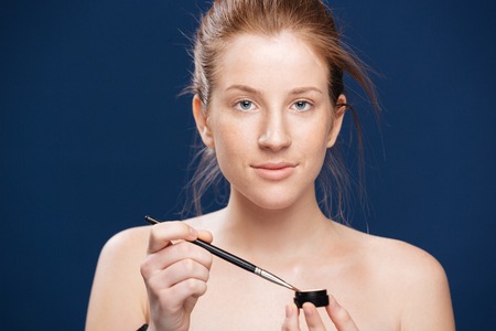 Beauty portrait of a young woman holding brush and cream over blue background