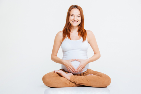 Pregnant woman sitting on the floor and showing heart gesture on her belly isolated on a white backgorundの写真素材