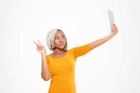 Playful cute young african american woman making selfie with tablet and showing victory sign over white backgroundの写真素材