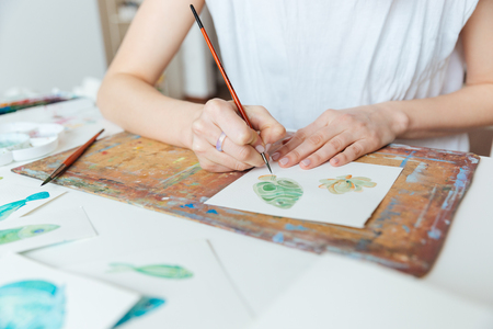 Closeup of hands of woman artist painting with paintbrush and watercolor paints on the tableの写真素材