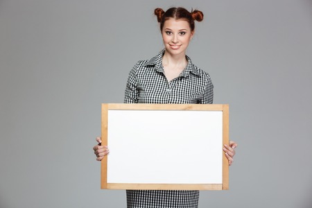 Cute lovely cheerful young woman standing and holding blank whiteboard over grey backgroundの写真素材