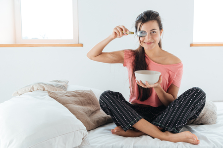 Happy playful young woman covered her eye with spoon and sitting on bed in bedroomの写真素材