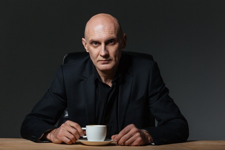Handsome businessman sitting at the table and drinking coffee over black background
