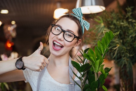 Happy charming young woman florist showing thumbs up in flower shopの写真素材