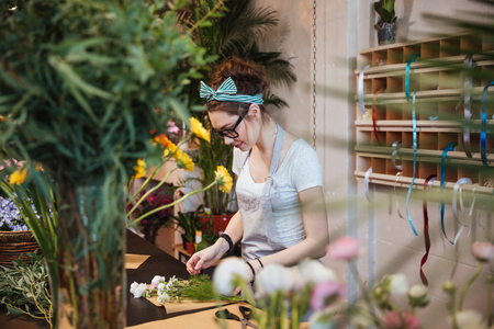Beautiful young woman florist in apron and glasses making bouquet and working in flower shopの写真素材