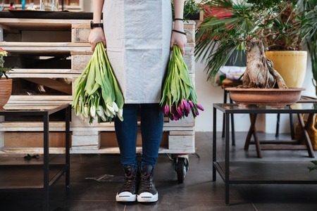 Closeup of young woman florist in sneakers standing and holding two bunches of tulips in flower shopの写真素材