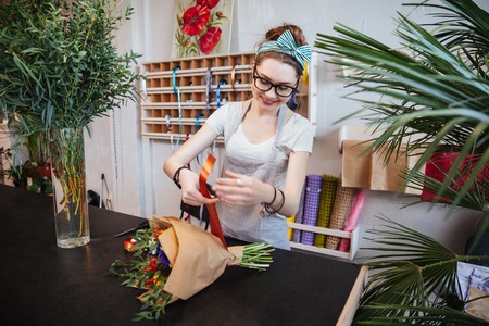 Smiling pretty young woman florist standing and making bouquet of flowers with red ribbon