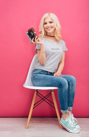 Happy young woman sitting on the chair and holding photo camera over pink backgroundの写真素材
