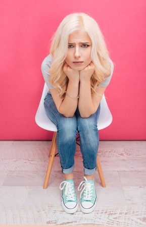 Unhappy blonde woman sitting on the chair and looking at camera over pink backgroundの写真素材