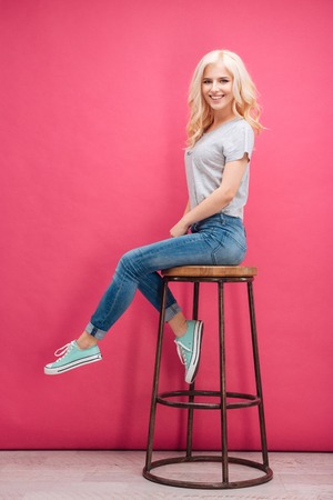 Full length portrait of a smiling blonde woman sitting on the chair over pink background and looking at cameraの写真素材