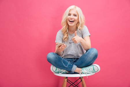 Cheerful woman listening music in headphones and showing thumb up over pink backgroundの写真素材