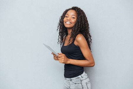 Smiling afro american woman standing with tablet computer over gray background and looking at cameraの写真素材