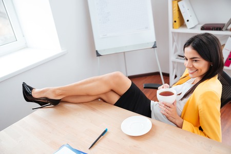 Happy businesswoman resting with legs on the table and drinking coffee in officeの写真素材