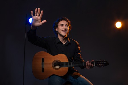 Smiling man holding guitar and waving hand over dark background