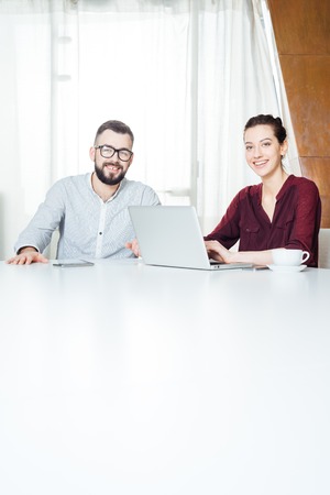 Portrait of two smiling attractive young businesspeople sitting at the table and using laptop in...