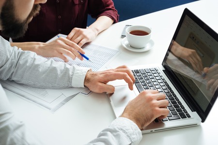 Closeup of hands of two young businesspeople typing on laptop and working together at white table