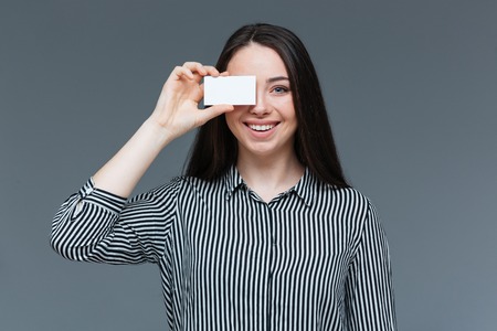 Happy woman covering eye with blank card over gray backgroundの写真素材