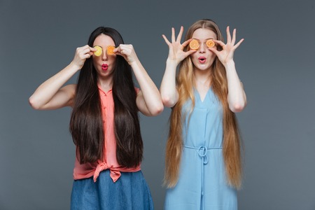 Two funny amazed cute young women with long hair covered their eyes with marmalade candies over grey...